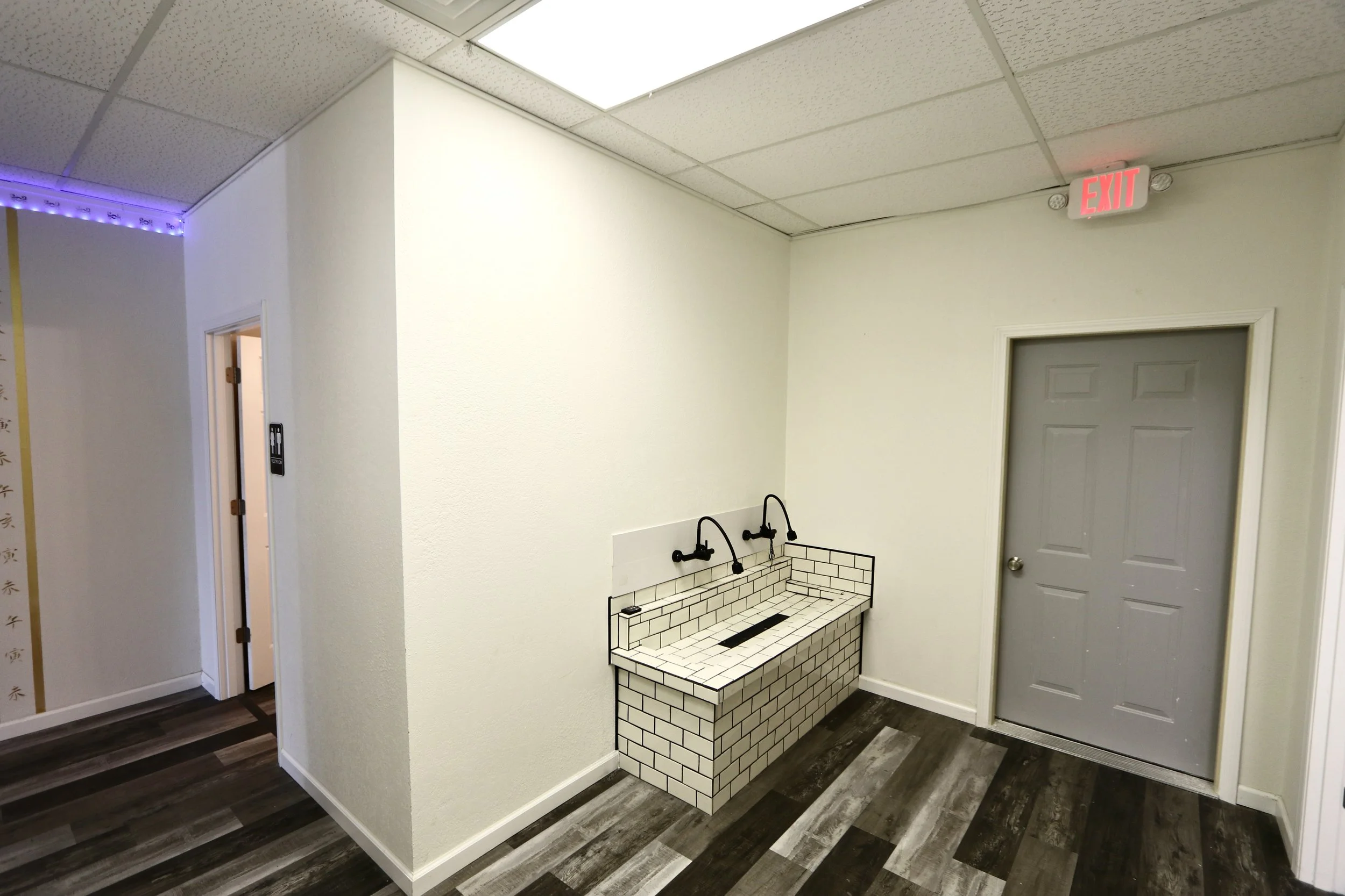 Indoor space with white walls, a gray door, a double black sink with white subway tile pattern, and a wood floor.