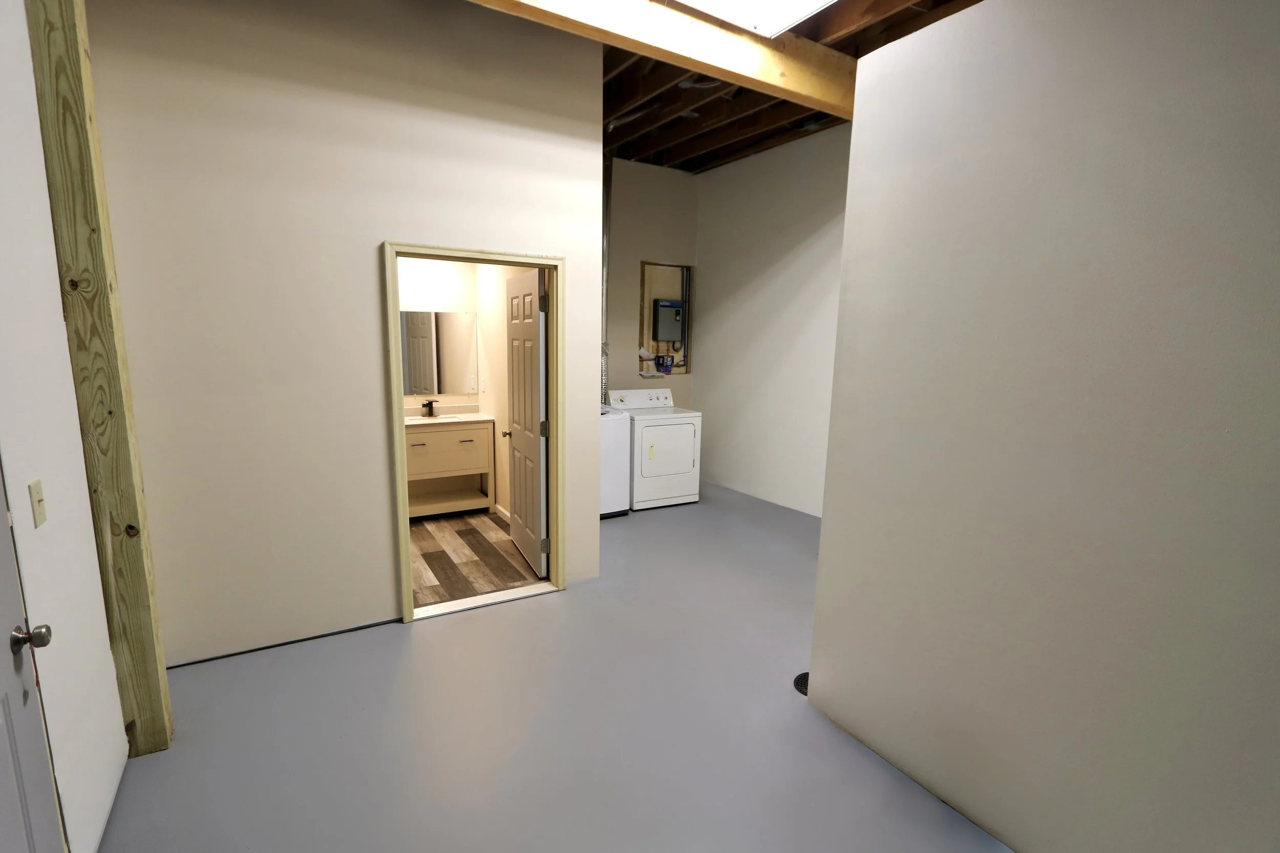 Empty laundry room with a washer and dryer, open doorway to a bathroom with a sink and mirror, visible drywall, and unfinished ceiling with exposed wooden beams.
