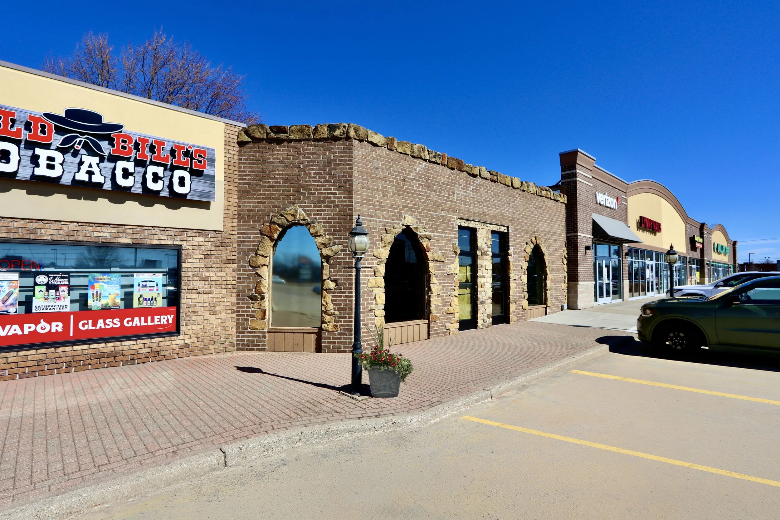 Strip mall with various retail stores, including Bill's Bacco, Vapor Glass Gallery, and Verizion, with parking lot and cars in front, lamp posts, flower planter, and blue sky.