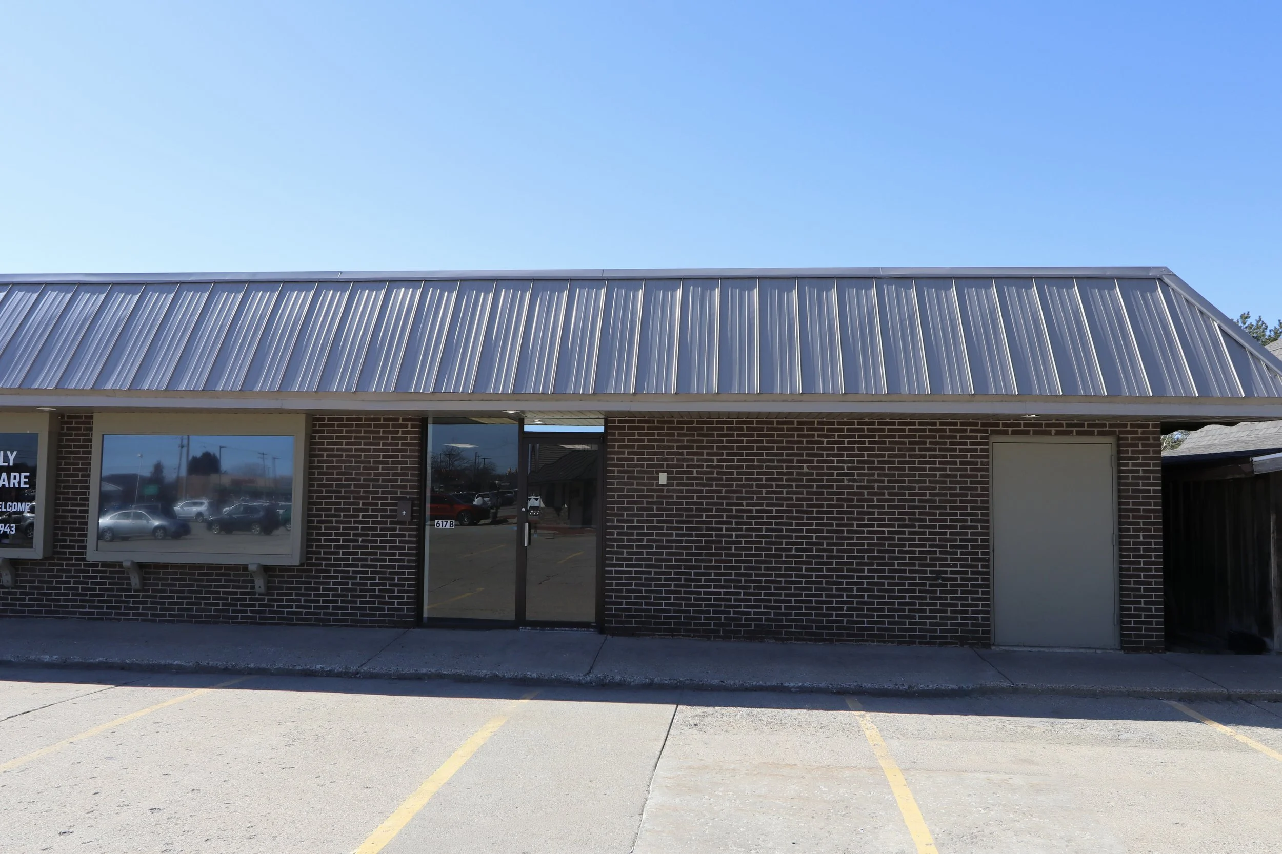 A small brick commercial building with a metal roof, glass front door, and parking spaces in front.