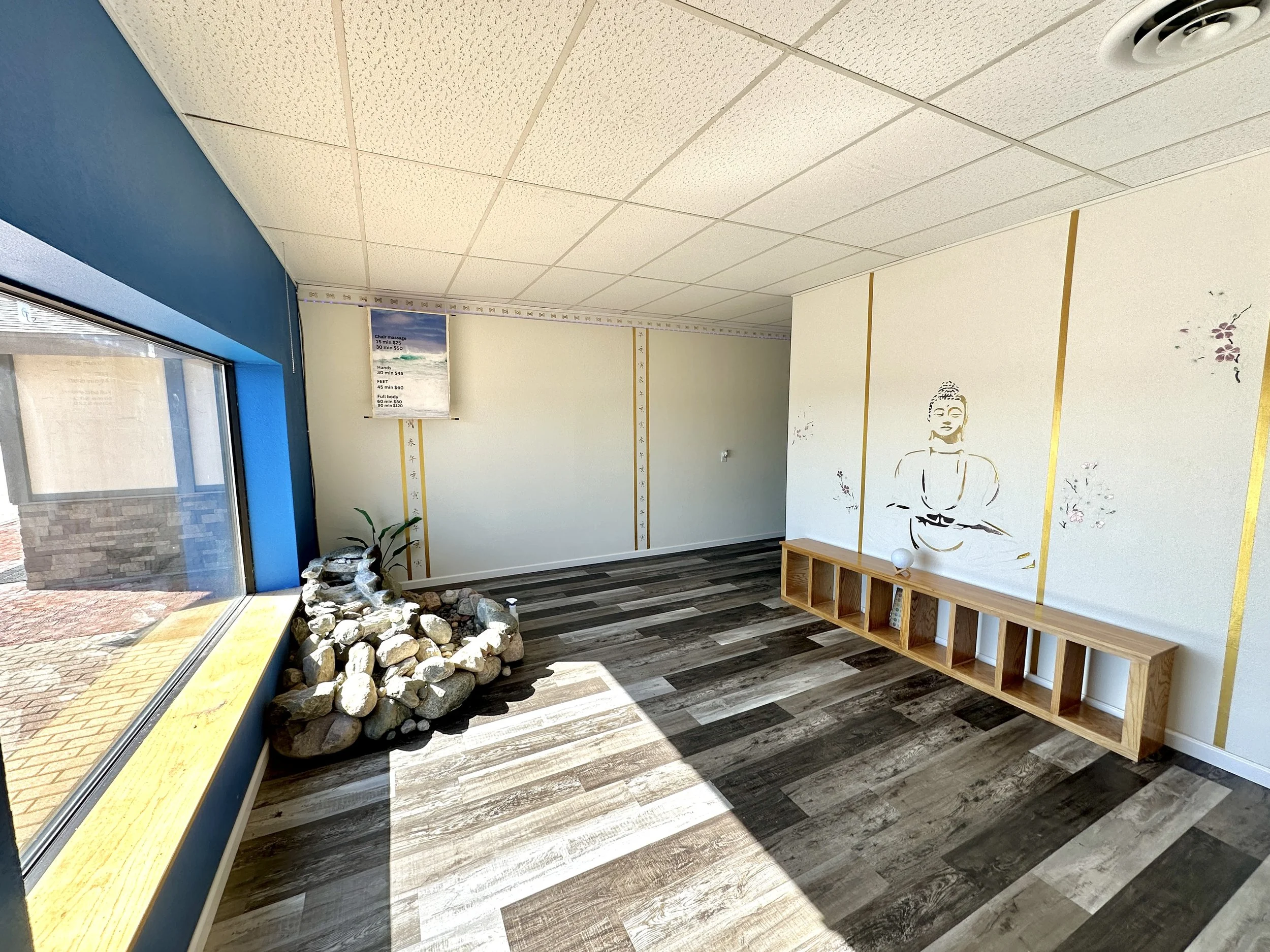 Empty room with wooden floor, tiled ceiling, a large window, a rock garden in the corner, a Buddhist mural on the wall, and a wooden shelf with a decorative ball.