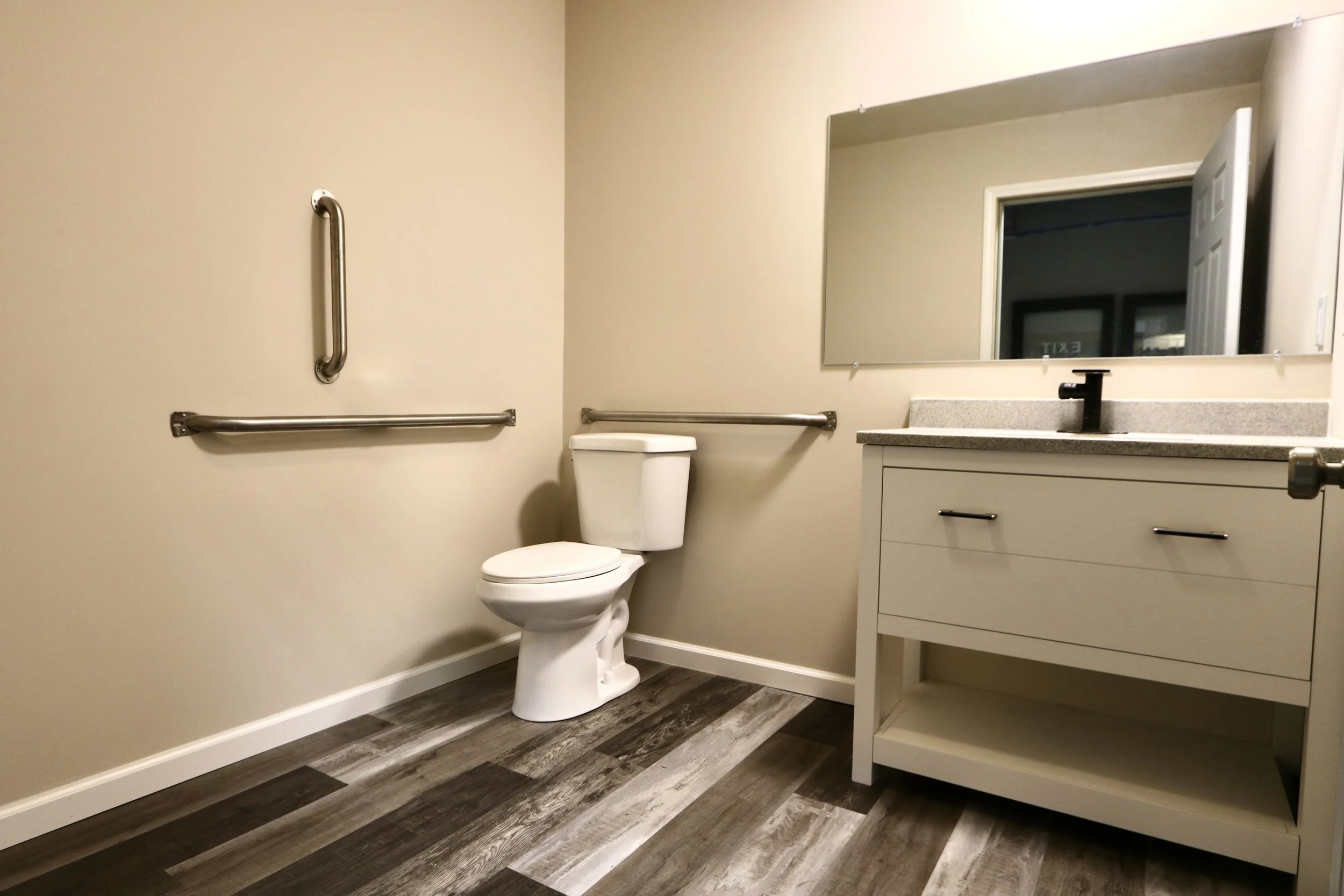 A bathroom with beige walls, a white toilet with grab bars on the wall behind it and beside it, a white vanity with a gray countertop, a black faucet, and a large rectangular mirror above the sink, all on a wood-patterned floor.