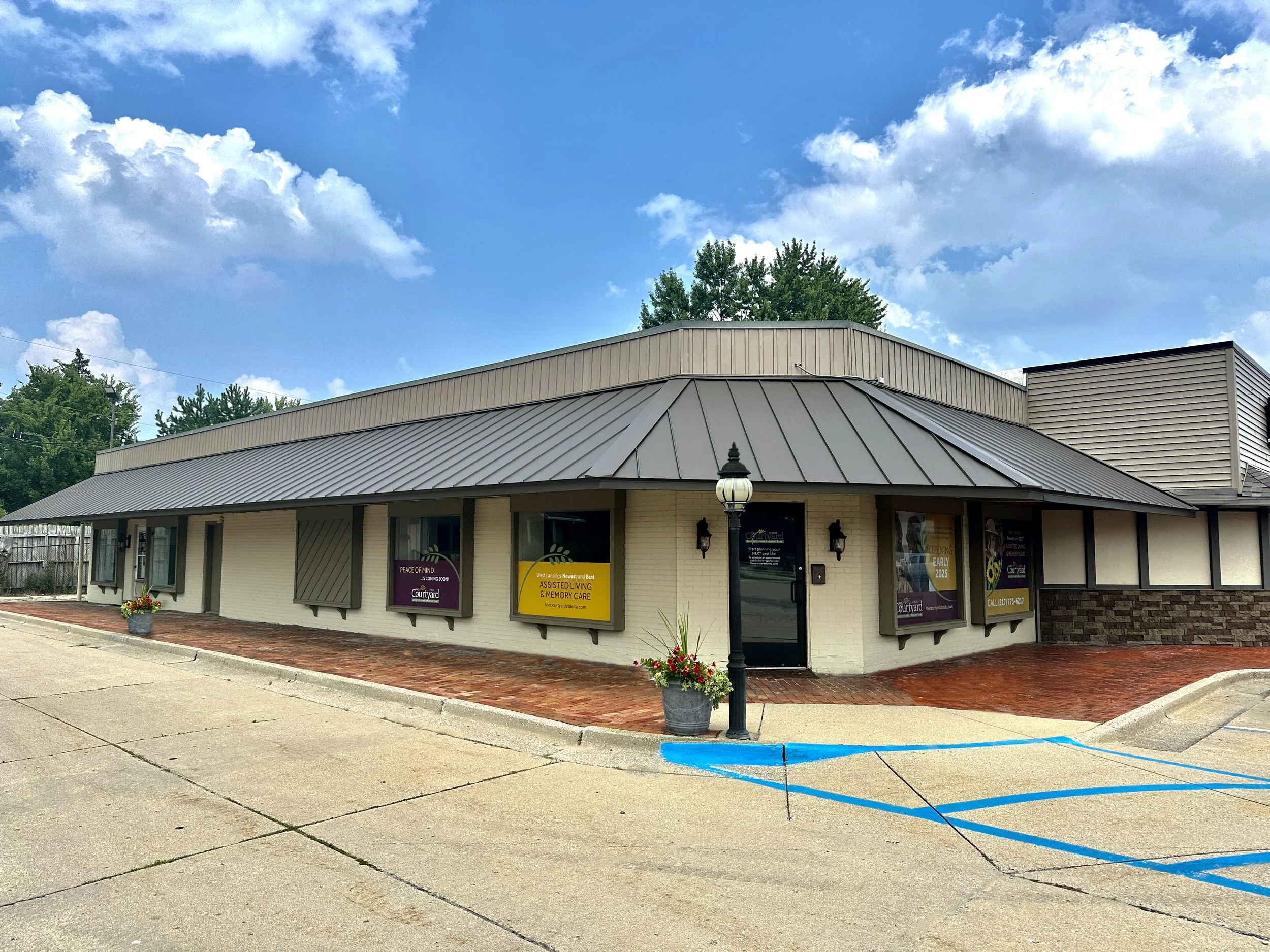 Exterior of a single-story building with a metal roof, multiple windows with signs, a lamp post, and a wheelchair accessible parking space marked in blue