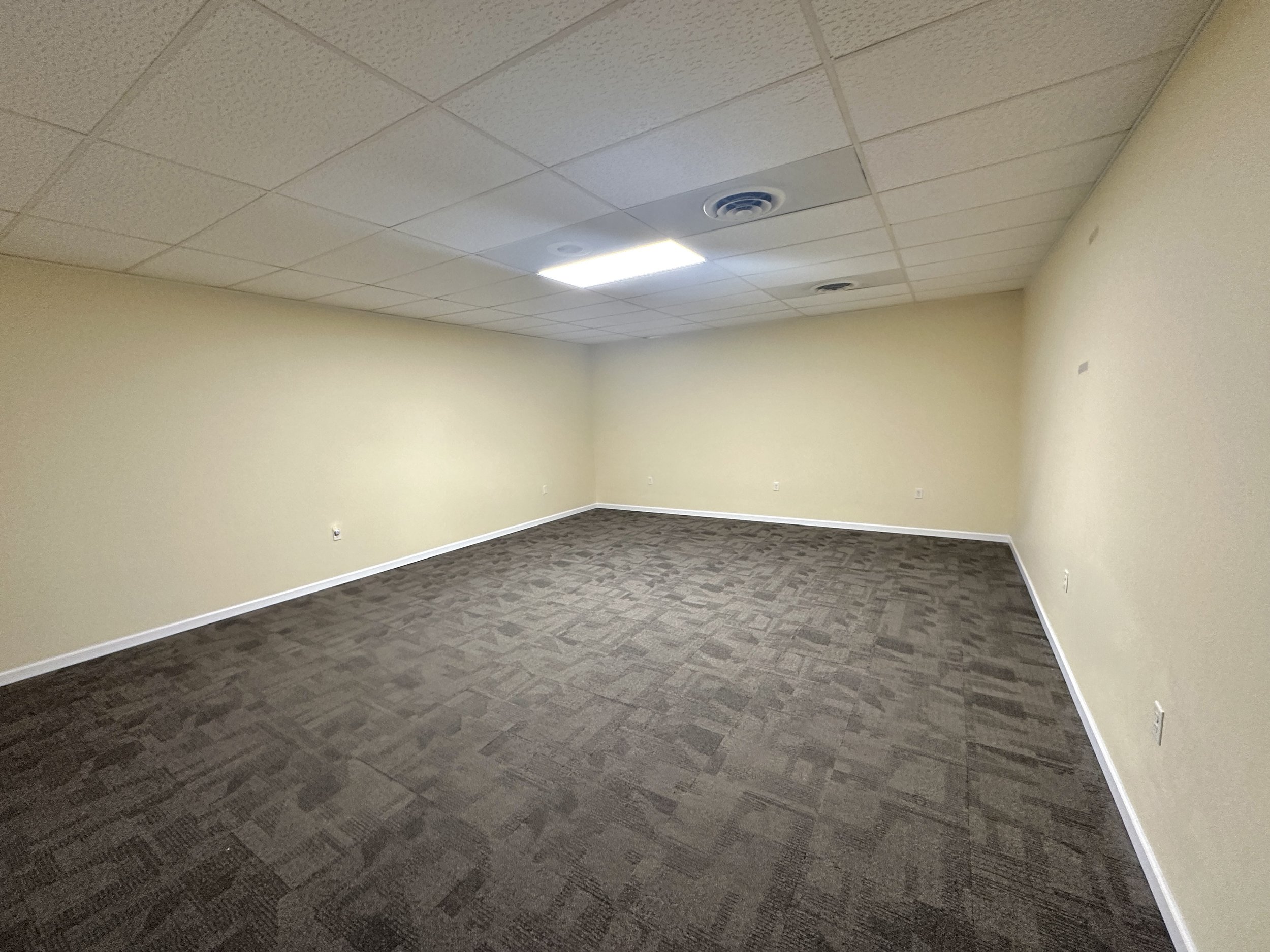 An empty rectangular office or meeting room with beige walls, a suspended ceiling with acoustic tiles, a central fluorescent light, and a dark patterned carpeted floor.