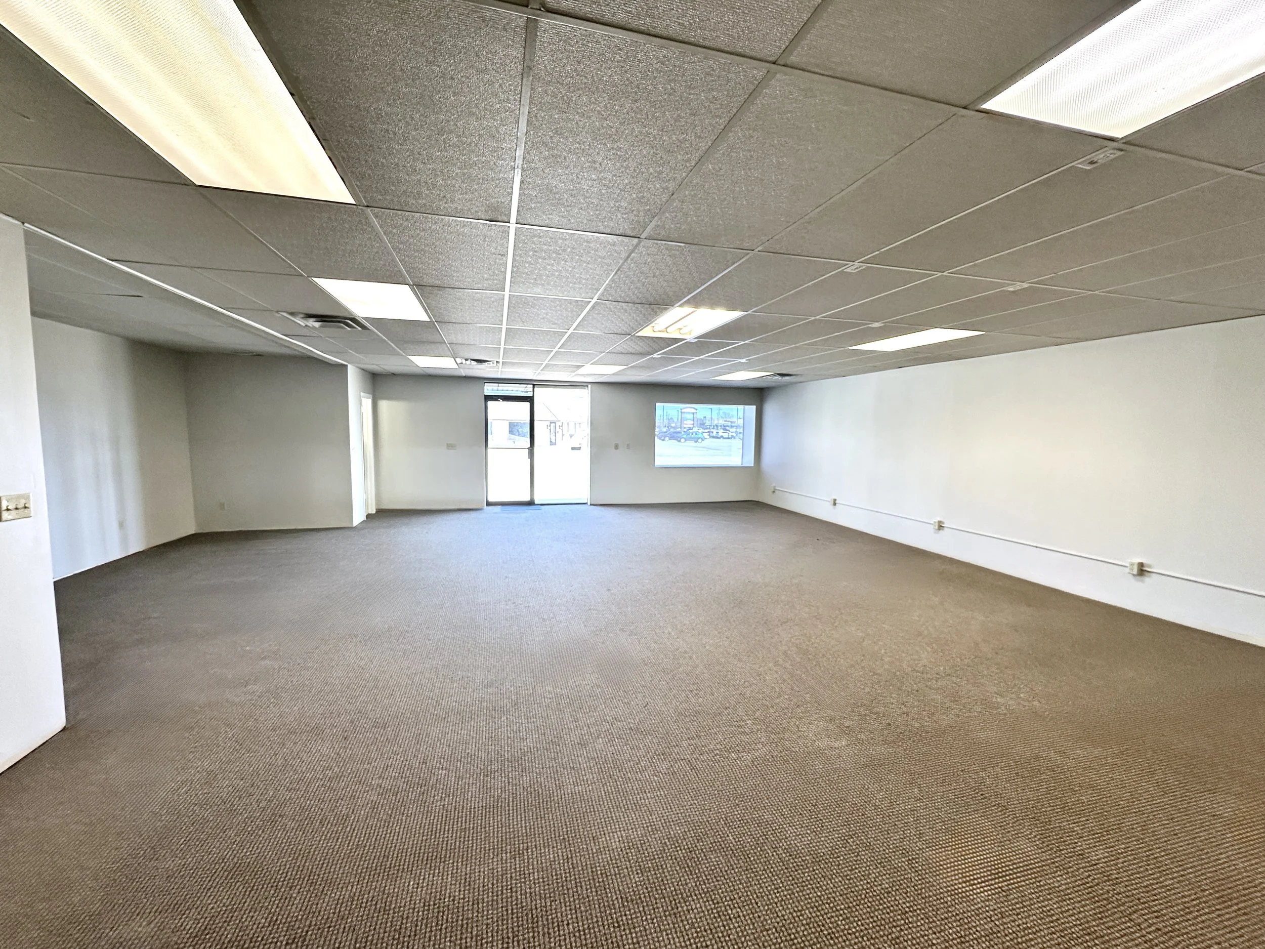 Empty commercial space with carpeted floor, white walls, and a suspended ceiling with fluorescent lighting fixtures. Large windows and a glass door at the far end provide natural light.