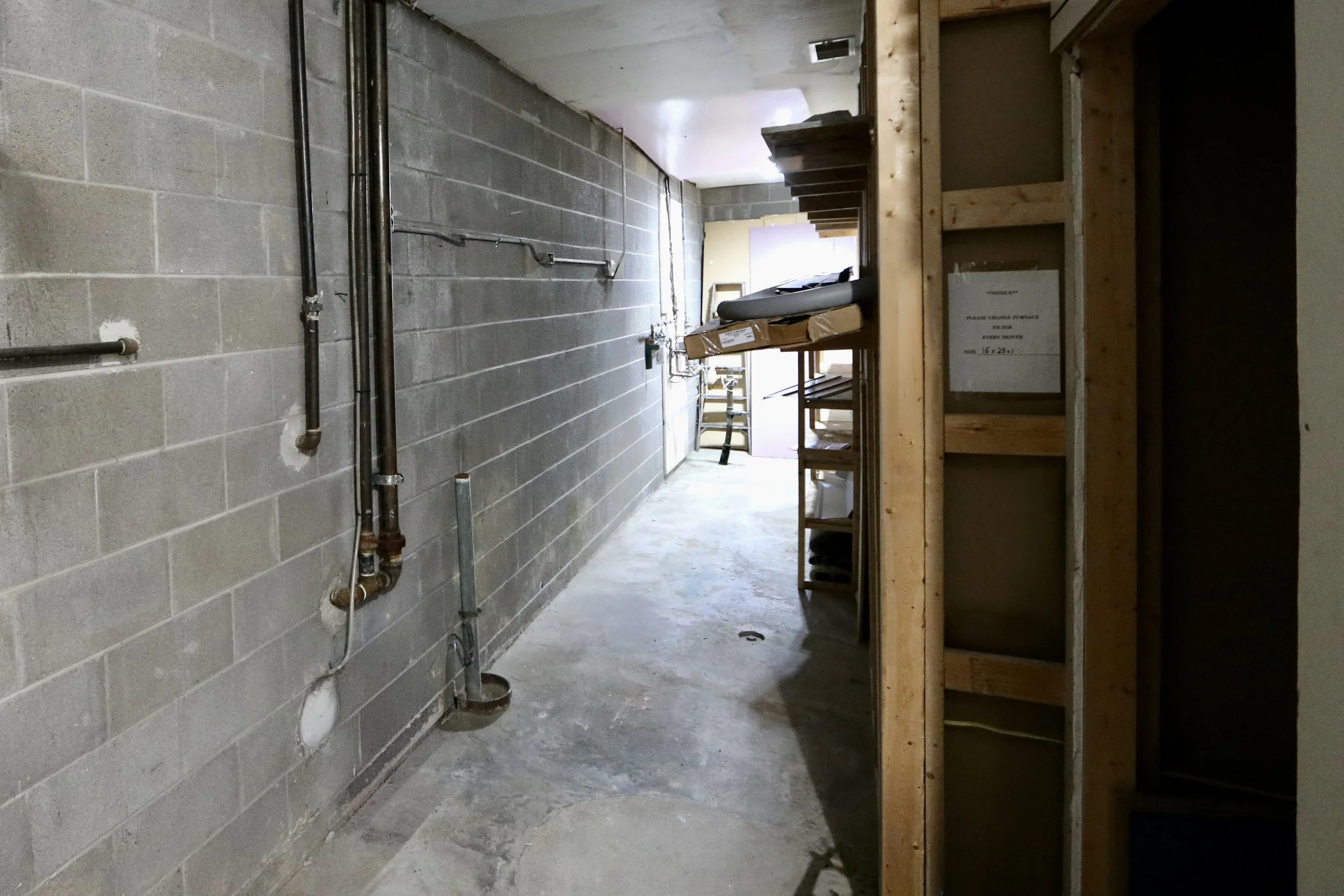 Construction storage area with cinder block walls, exposed pipes, wooden shelving, and a ladder at the end of the corridor.