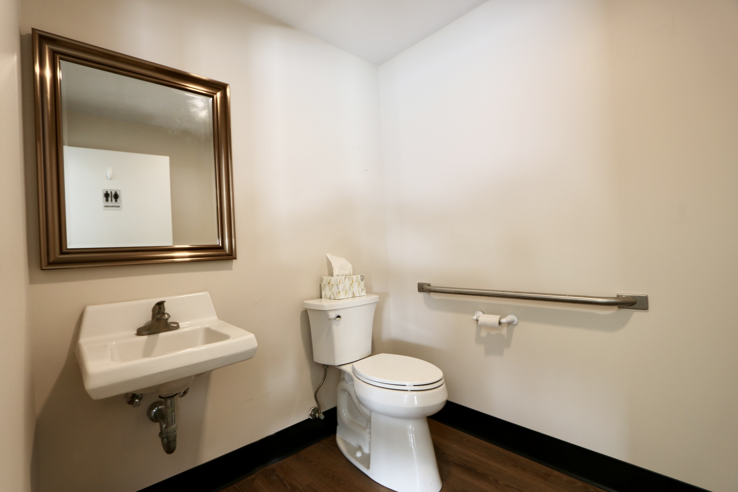 A small bathroom with a wall-mounted sink, a mirror with a bronze frame, a toilet with tissues on top, a horizontal grab bar, and a toilet paper holder on the wall.