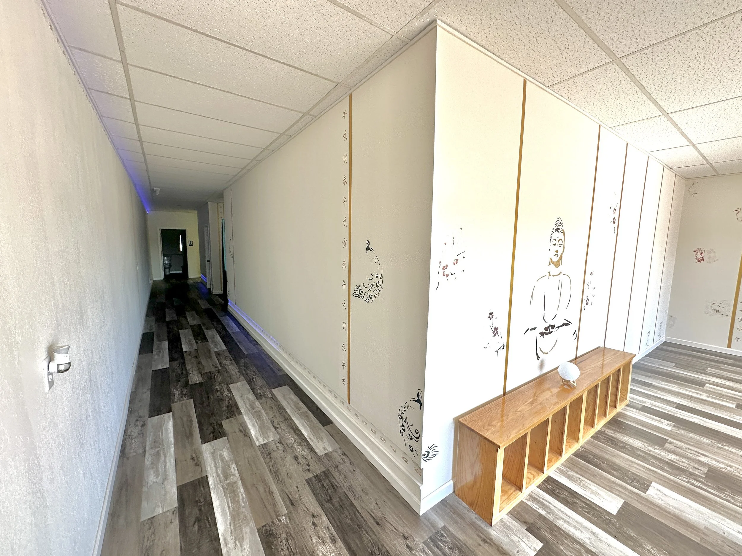 A hallway with a white wall decorated with Asian-style artwork, including a peacock and a figure in traditional attire. The floor has a striped, wood-like pattern, and there is a wooden shoe rack against the wall. The ceiling has a standard drop ceil