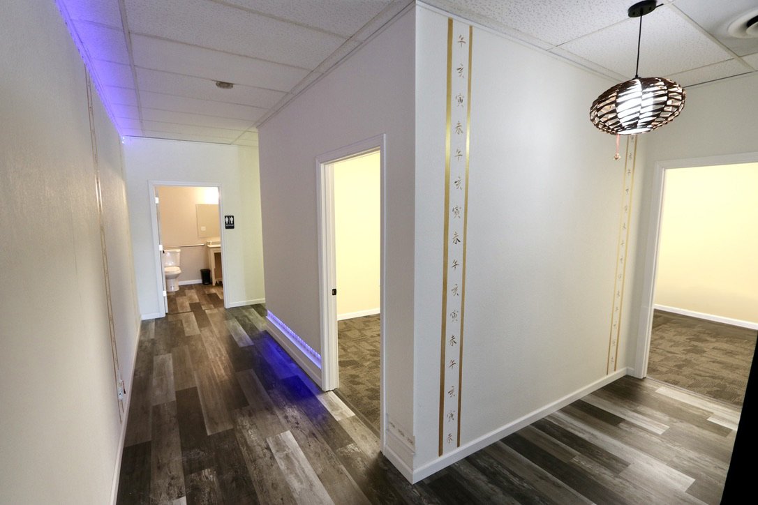 Interior hallway with wood flooring, leading to an open bathroom and several rooms, featuring white walls and decorative vertical gold lines with Asian script, a hanging pendant light, and purple accent lighting on the wall.