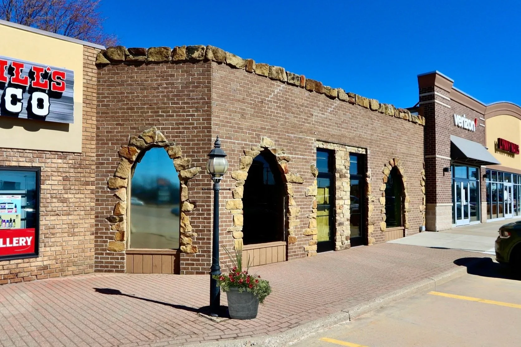 Image of a commercial building with brick walls, arched windows framed with stone, a lamp post, and storefront signs for Verizon and H Mart. The sky is clear and blue.