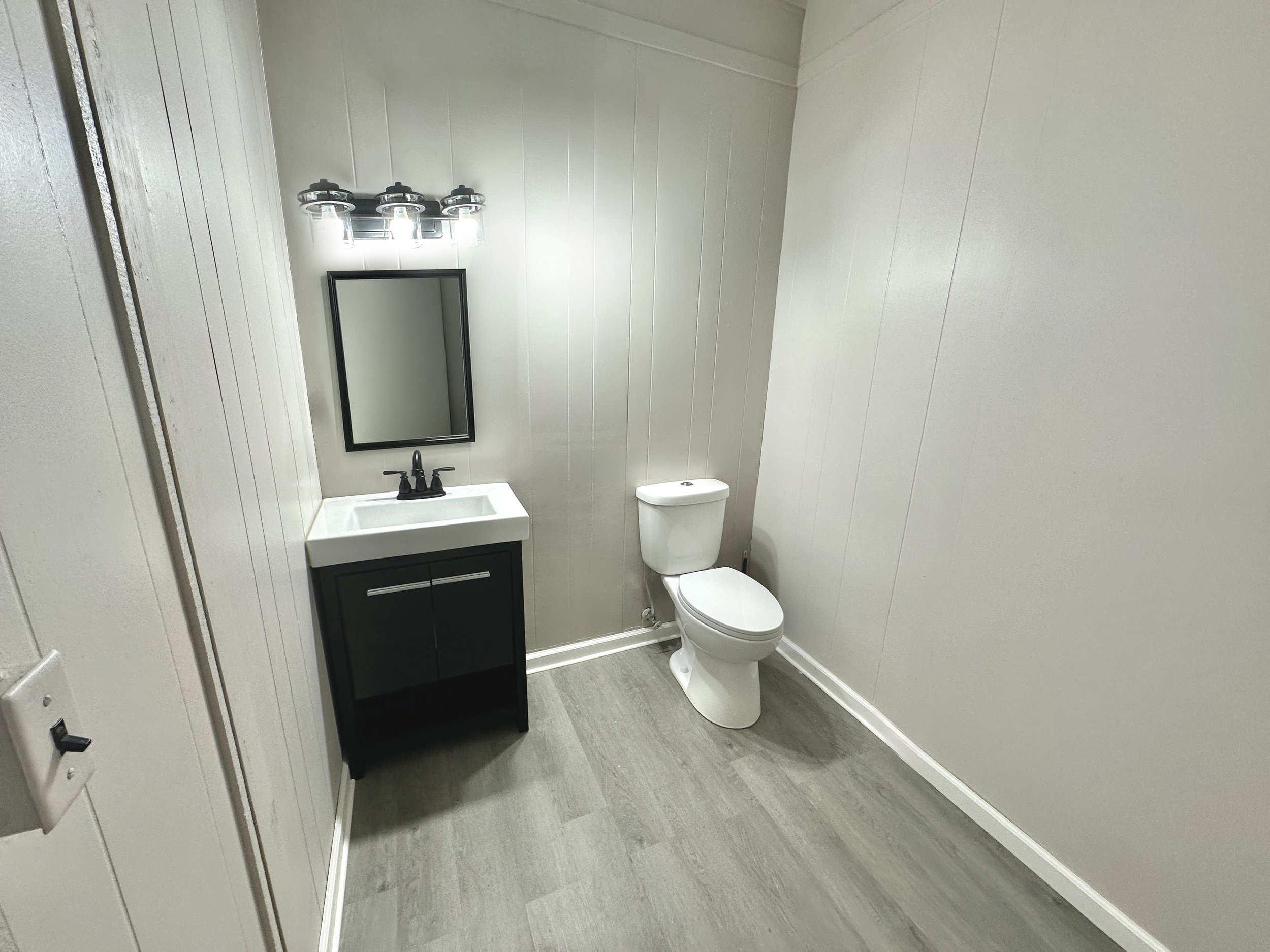 A small bathroom with light-colored paneled walls, a white toilet, a small black vanity with a white sink, and a rectangular mirror above the sink. A modern light fixture with multiple bulbs is above the mirror. The floor is light gray vinyl or lamin