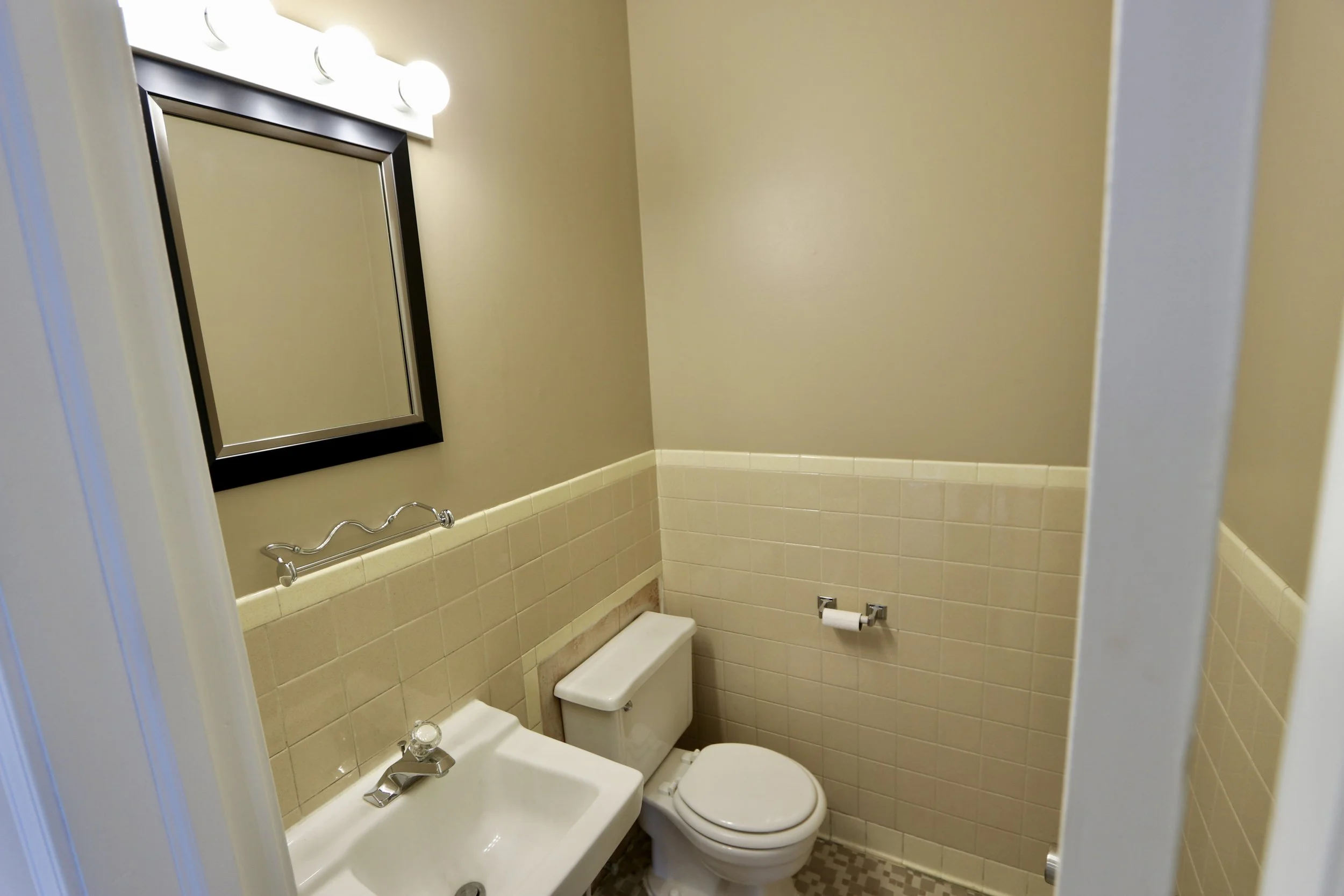 Small bathroom with beige tile halfway up the walls, a white toilet, a small white sink, a black-framed mirror, and a three-bulb light fixture above the mirror.
