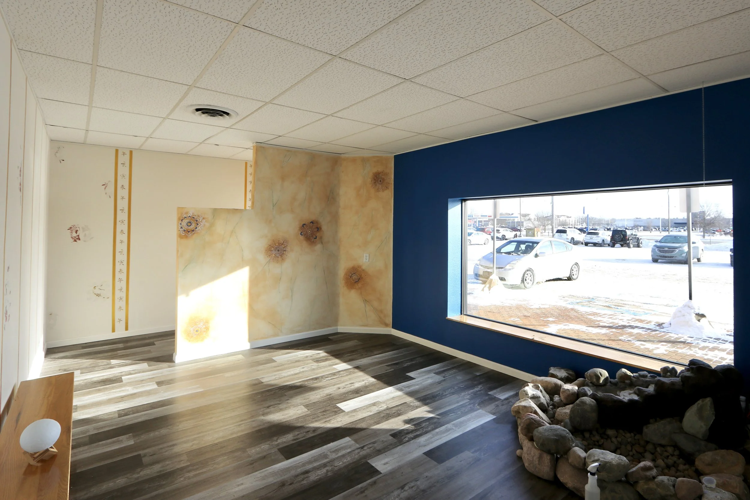 Empty interior room with wooden floor, large window showing snow-covered parking lot, painted walls with Asian-inspired designs, rock garden in corner, and ceiling tiles.