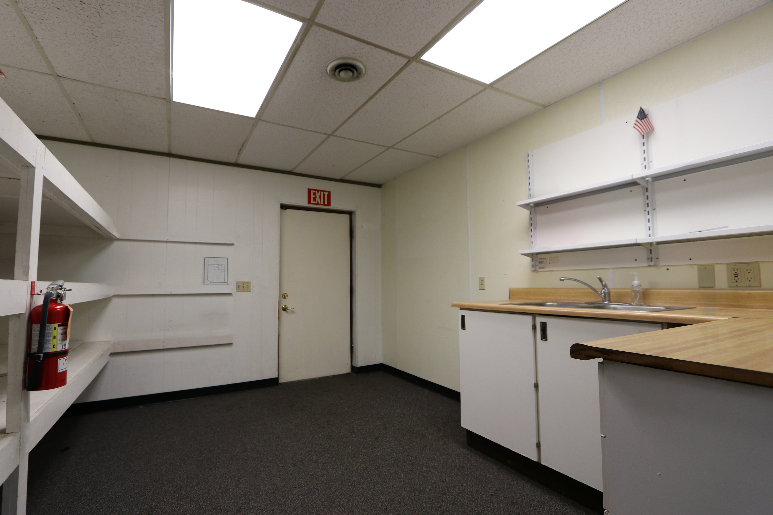Empty room with white walls and ceiling tiles, a sink with soap dispenser, open white shelves, and a closed door with an exit sign above.