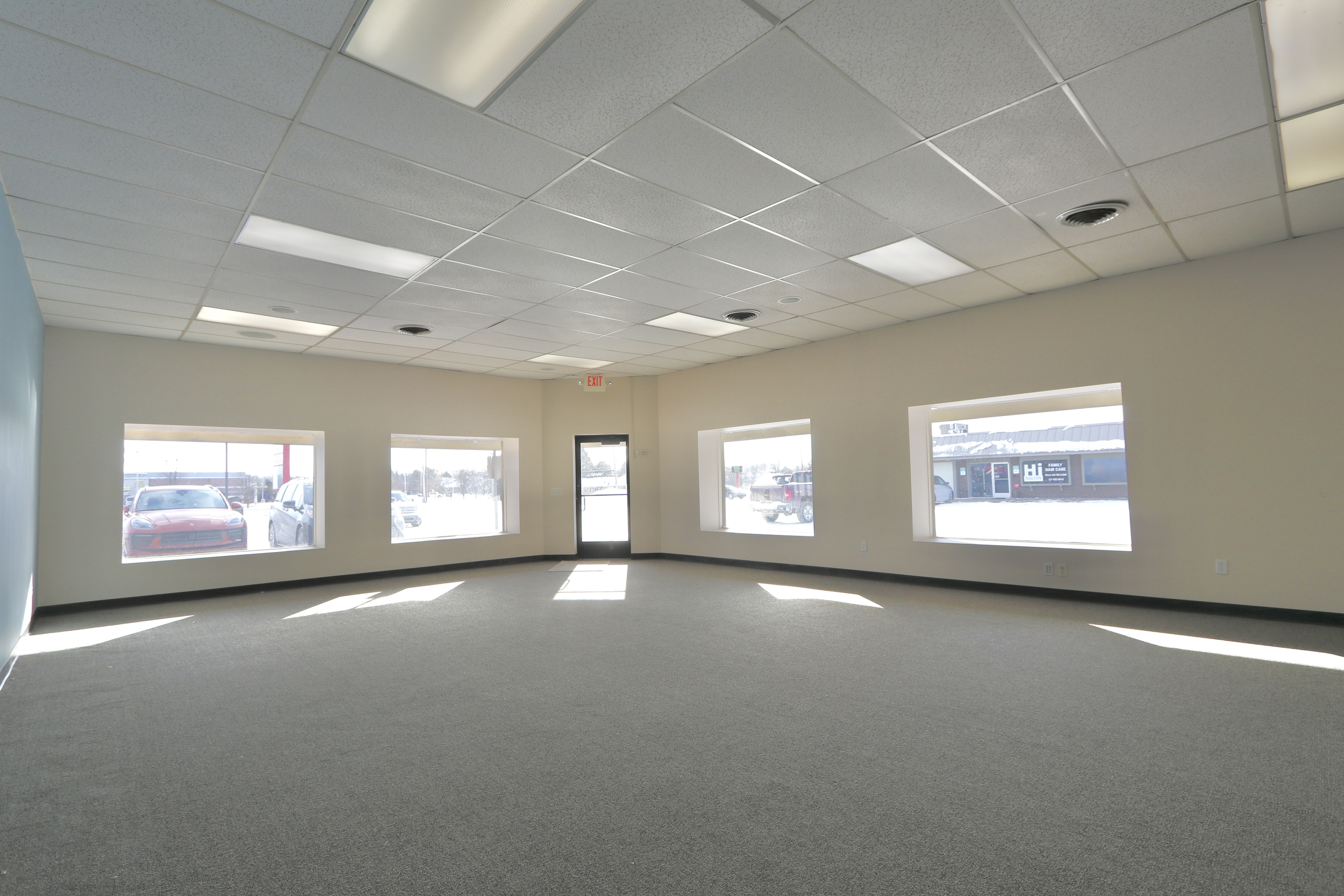 Empty commercial space with large windows, gray carpet, and a ceiling with fluorescent lighting.