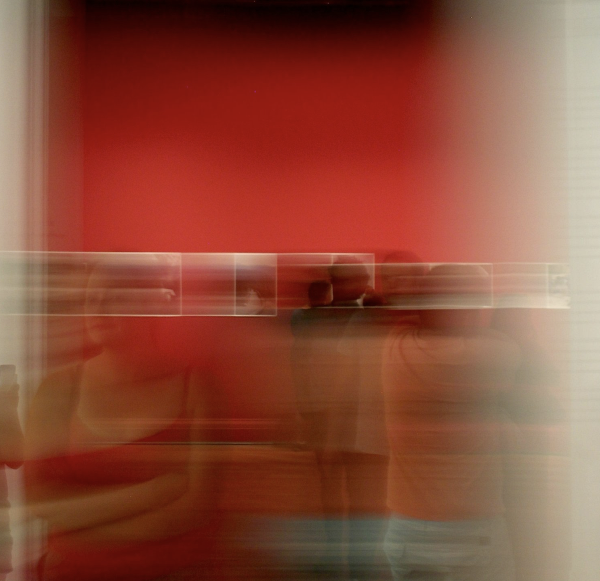 Blurred reflection of people in a mirror against a red wall.
