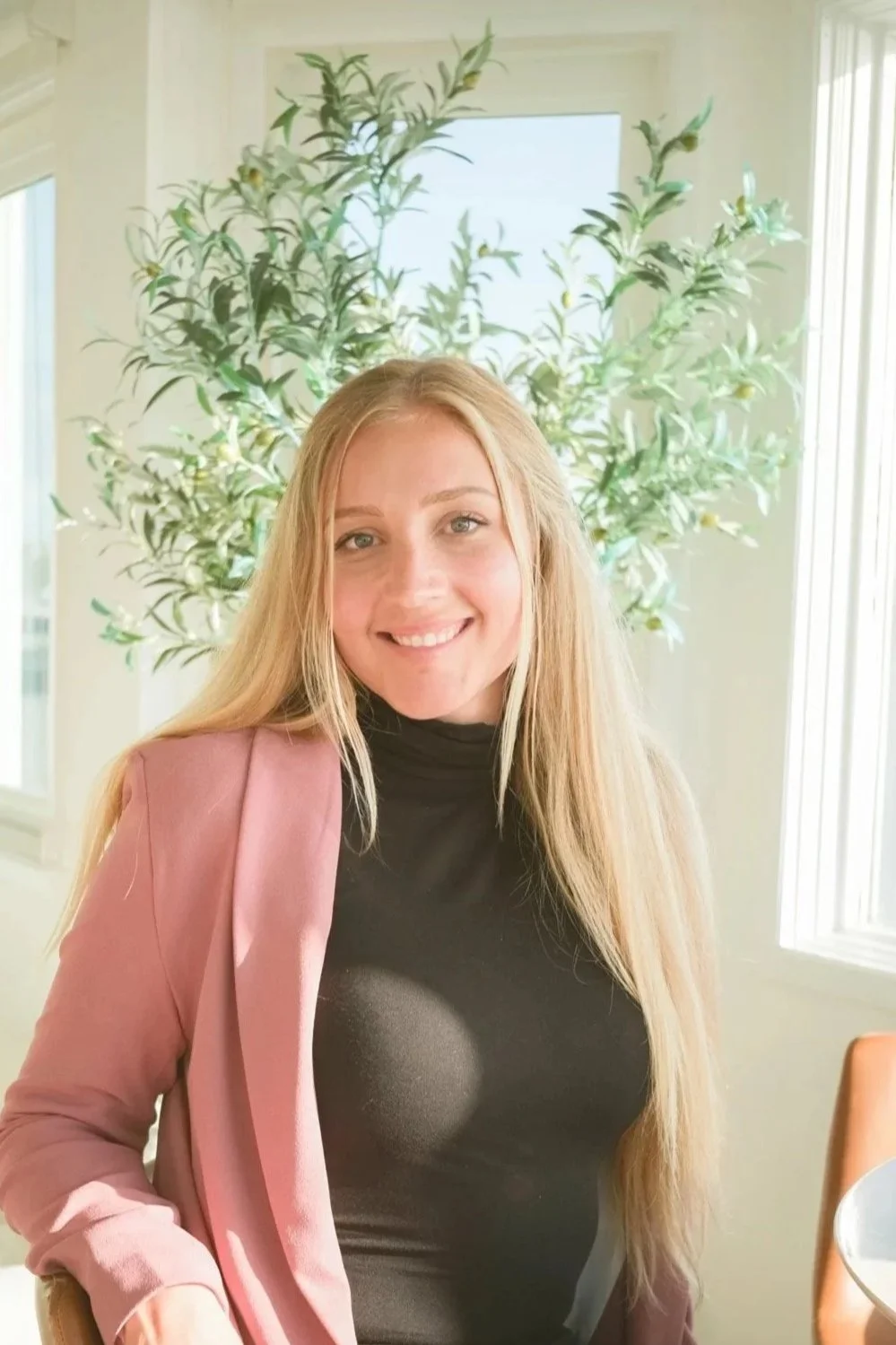 A young woman with long blonde hair wearing a black turtleneck and pink blazer, smiling at the camera, sitting in a sunlit room with a large green leafy plant and white windows in the background.