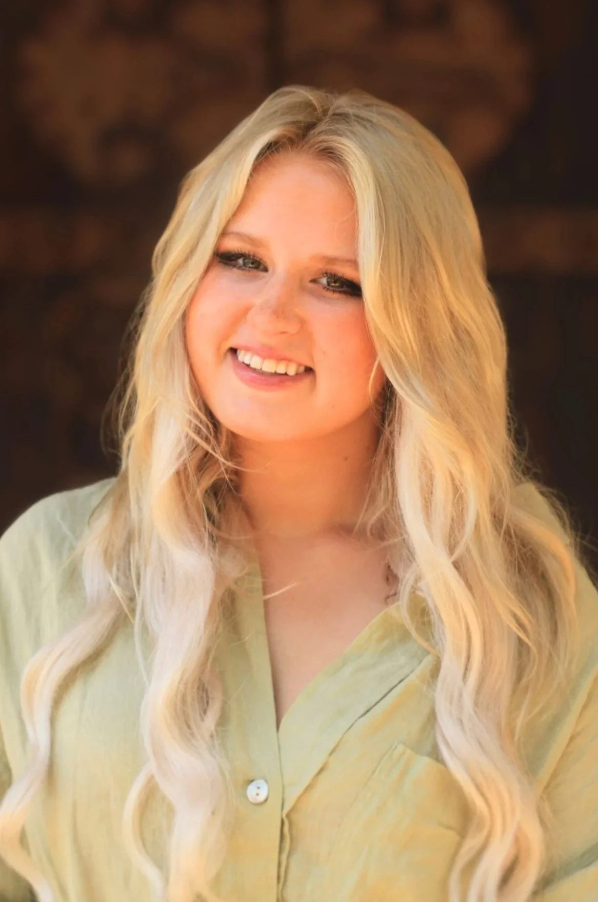 Young woman with long, wavy blonde hair, smiling, wearing a light yellow shirt, with a blurred dark background.