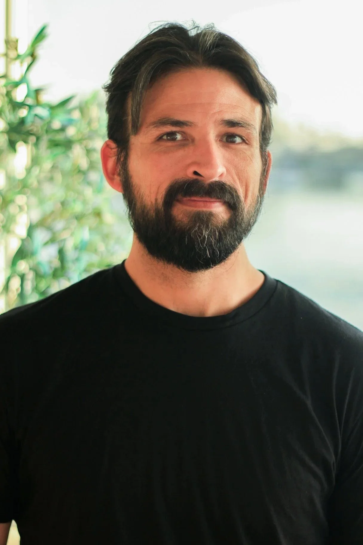 A man with a beard and dark hair, wearing a black t-shirt, standing outdoors near water and greenery.