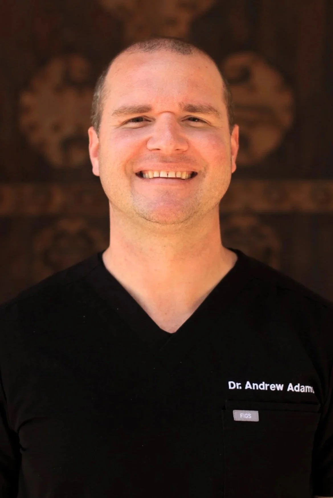 A smiling man wearing black medical scrubs with the name tag 'Dr. Andrew Adams' and 'FIGS' logo, standing outdoors with a dark, textured background.