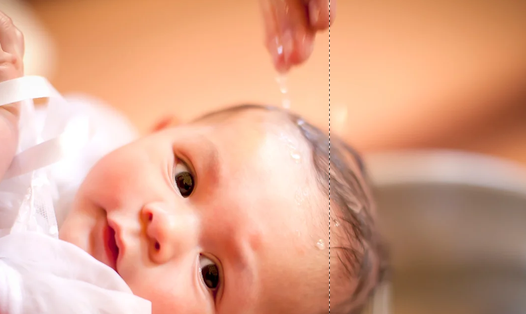 Close-up of a baby with water being poured onto their forehead from a person's hand.