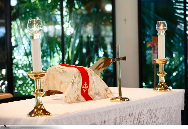 Eucharistic table setup with two lit candles, a chalice, a missal, and a white cloth with red and gold accents in a church or chapel setting.