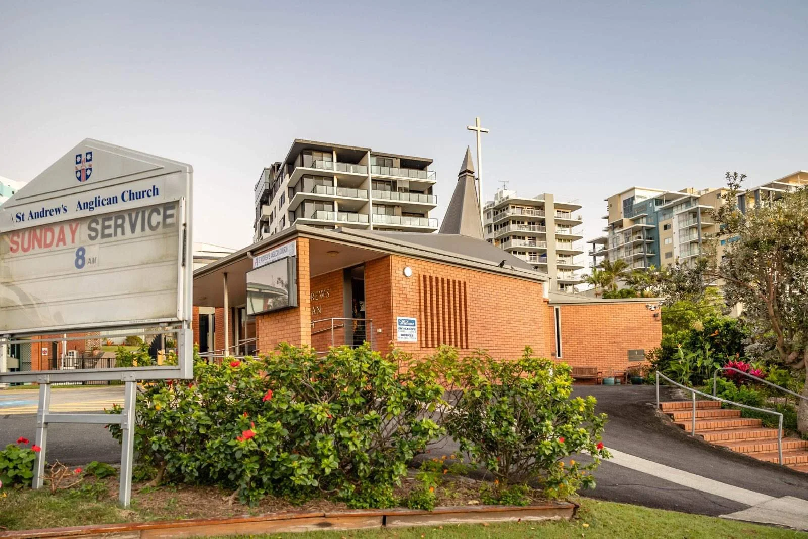 Exterior of St. Andrew's Anglican Church with a brick building, a small steeple with a cross, surrounded by greenery and multi-story residential apartments in the background.