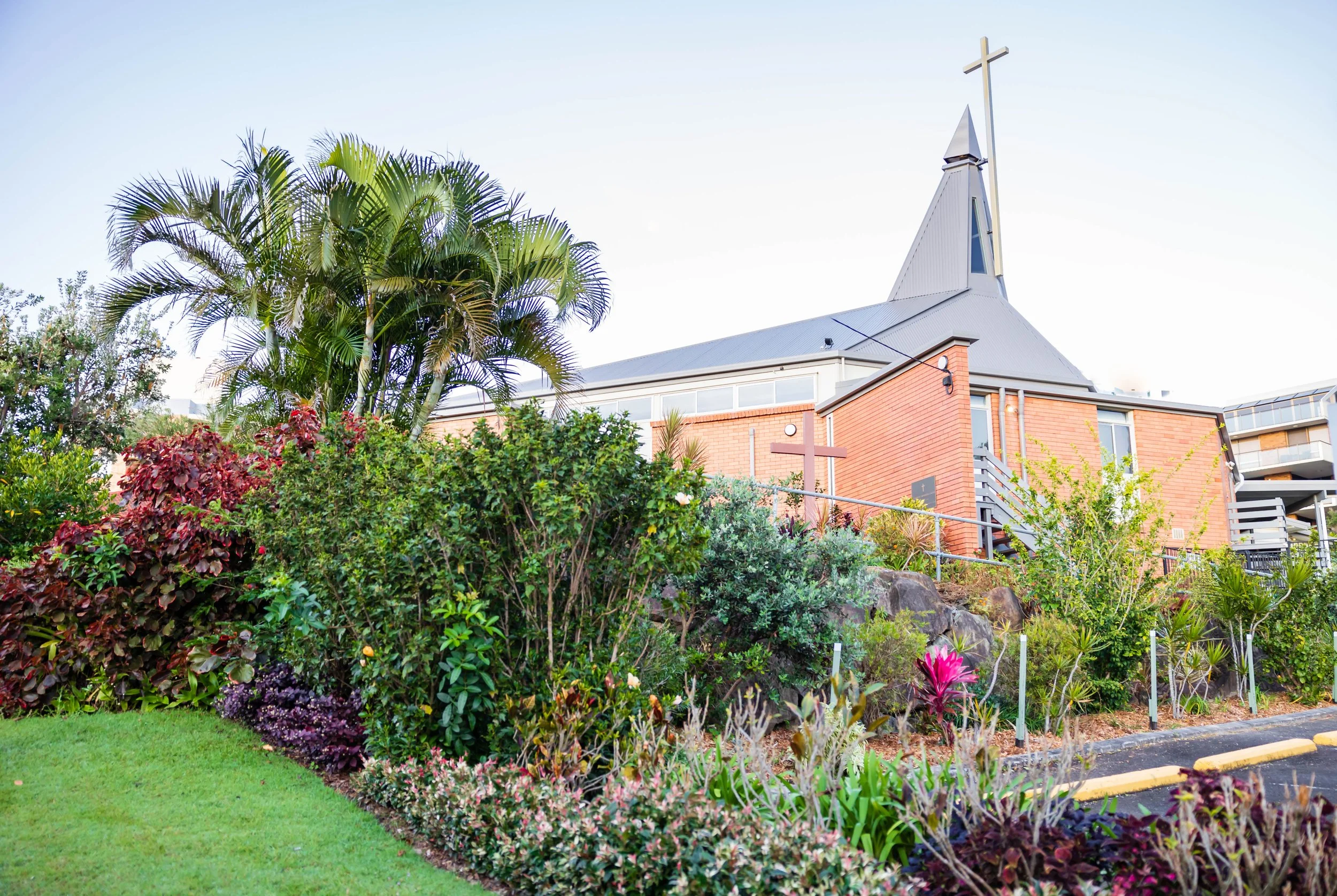 A church with a steeple topped by a cross, surrounded by lush green bushes, palm trees, and flowering plants.