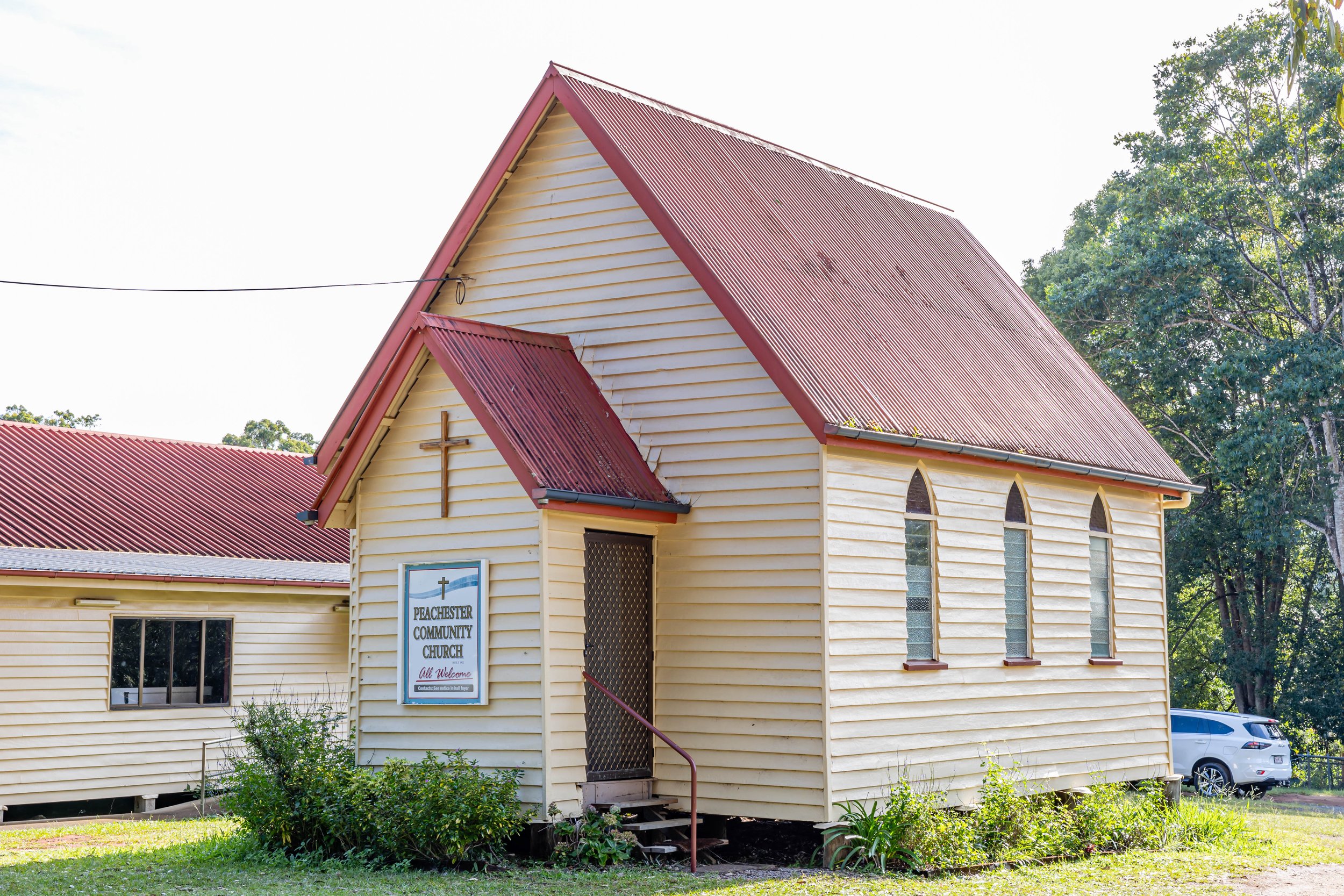 Yellow wooden church with red metal roof, small cross on front, and sign that reads 'Peachester Community Church, All Welcome', surrounded by green grass and bushes, with trees in the background.