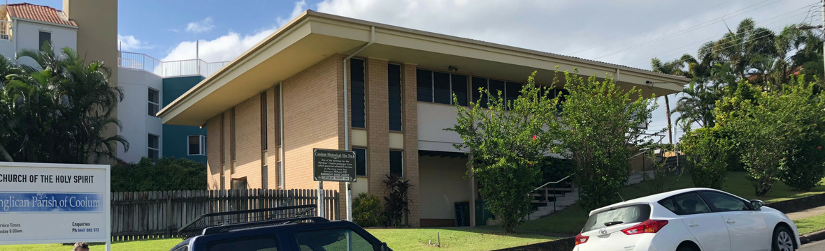 A church building next to a parking lot with a white car and a black vehicle parked outside, surrounded by trees and a blue sky.