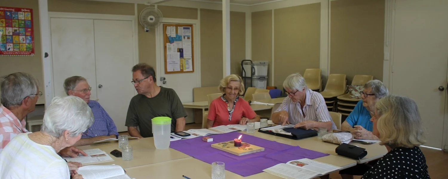 Group of elderly people sitting around a table in a meeting room, engaging in discussion with open books and notebooks, a purple cloth with a lit candle in the center on the table.
