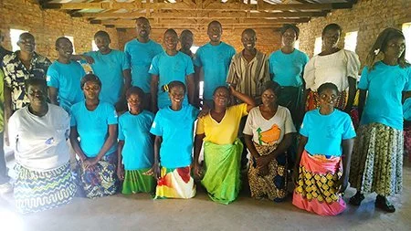 Group of women and girls in colorful clothing and blue shirts, posing indoors against a brick wall.
