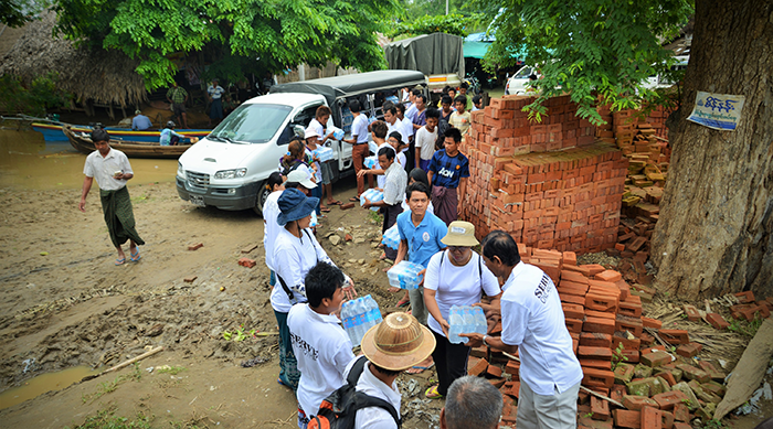 People in a line distributing water bottles outdoors near a tree and brick wall.