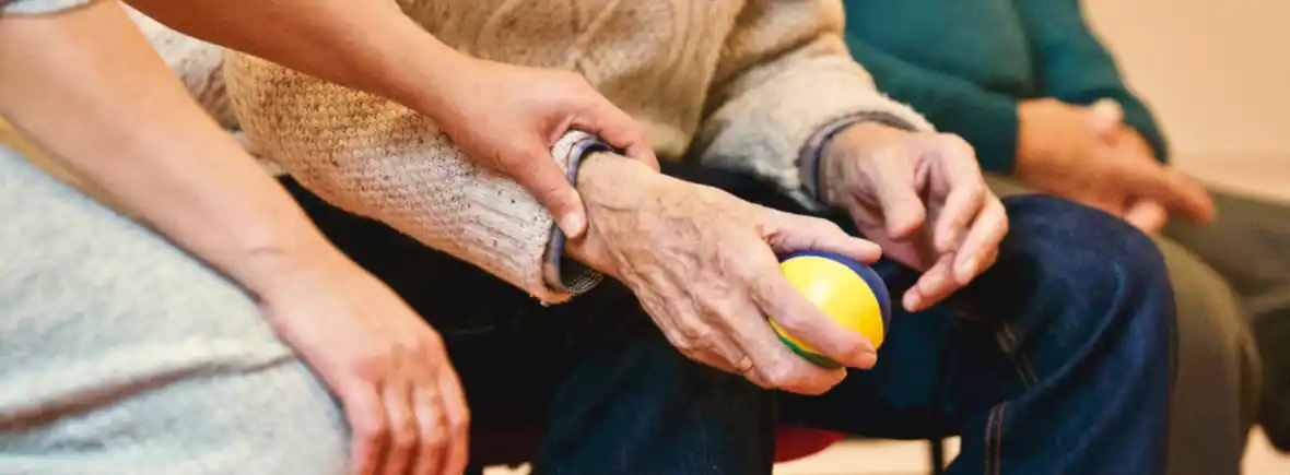 Close-up of elderly person holding a yellow and green ball with assistance from a caregiver's hand.