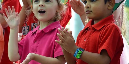 Two children, one girl in a pink shirt and one boy in a red shirt, clapping and participating in an outdoor activity.