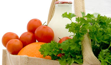 A grocery bag containing oranges, tomatoes, a bottle of milk, and fresh green cilantro.