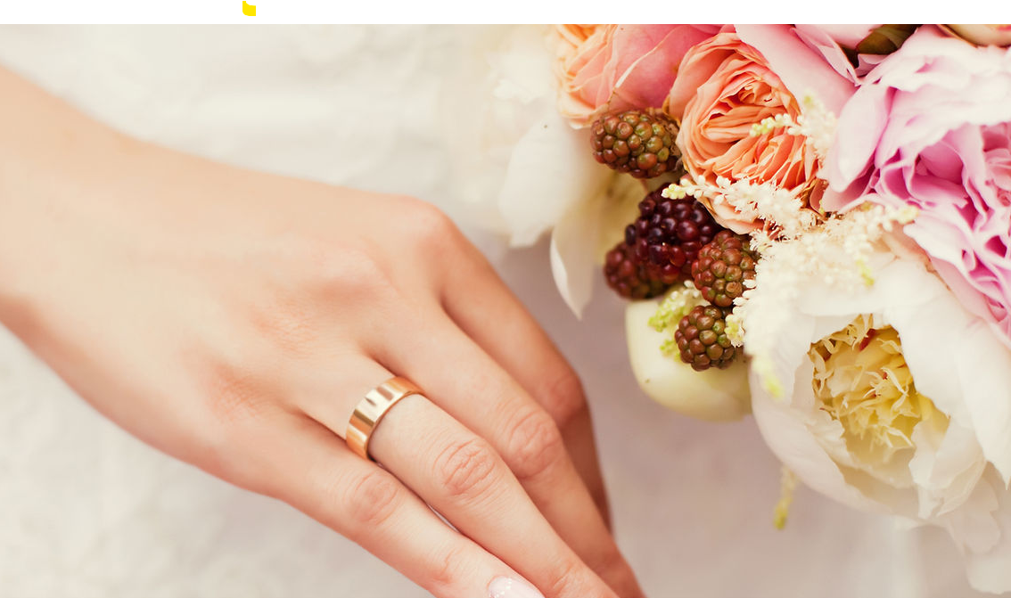 Close-up of a woman's hand wearing a gold wedding band, resting near a bouquet of pink and white flowers with some small berries.
