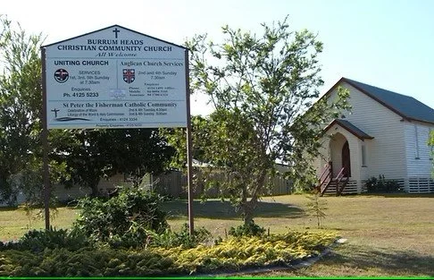 A church building and a large signboard displaying church information in a grassy area with some bushes and trees.