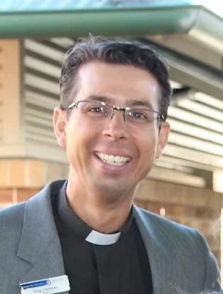 A man wearing glasses and a clerical collar, smiling outside a building.