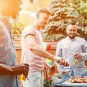 A group of people grilling and socializing outdoors with trees and sunlight in the background.