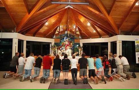 A group of people kneeling in prayer inside a church with a wooden ceiling and an altar at the front.