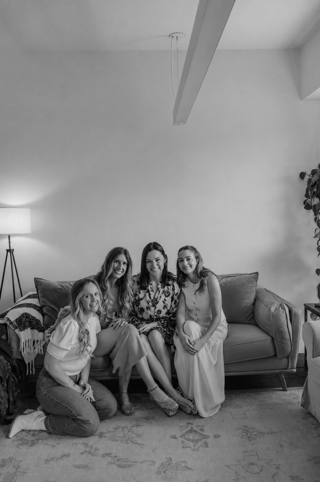 Four women sitting and kneeling on a sofa in a living room, smiling at the camera. The room has a lamp, a blanket, and a potted plant.