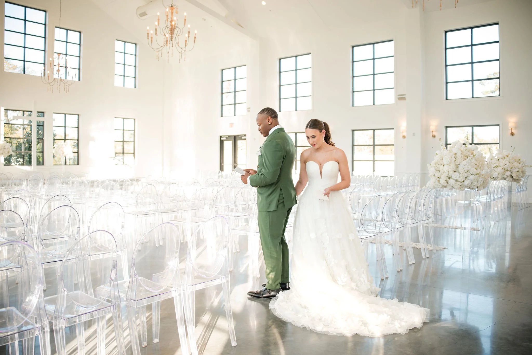 A bride and groom standing together in an empty, brightly lit wedding venue with rows of transparent chairs and large floral arrangements.