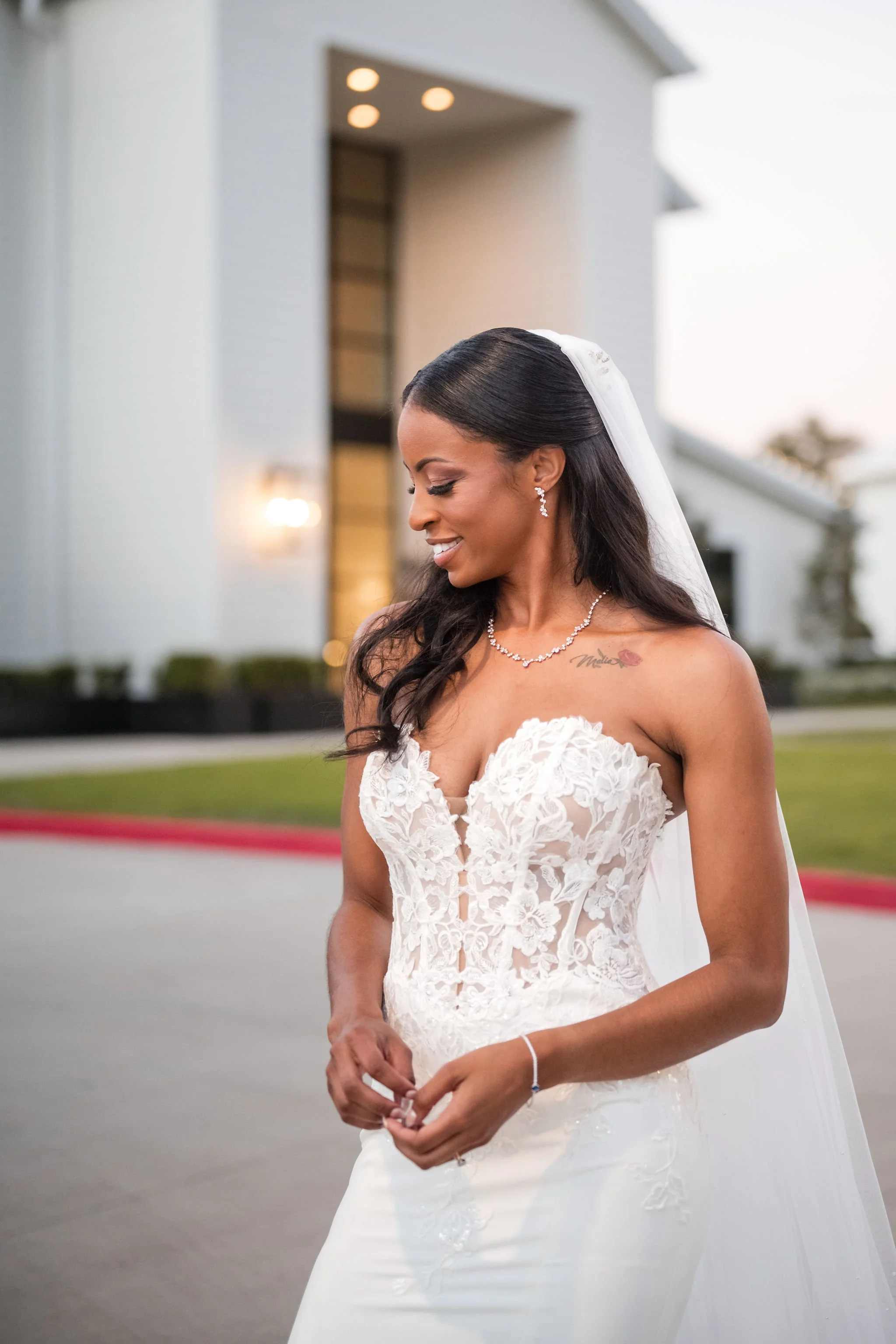 A bride in a white strapless wedding dress with lace details, standing outdoors in front of a modern building, wearing a veil and jewelry, smiling with her eyes closed.
