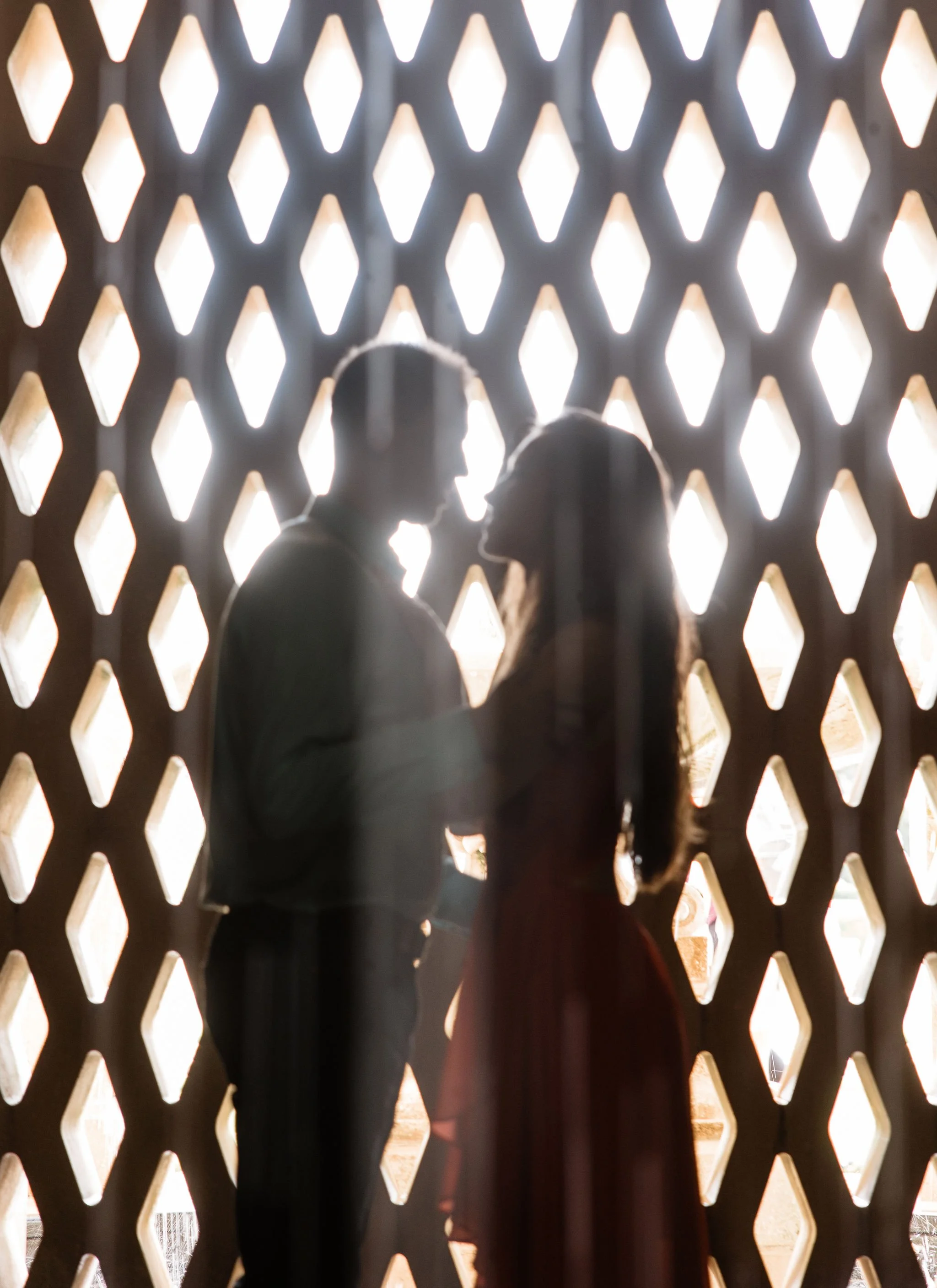 A silhouette of a couple facing each other closely in front of a decorative wall with diamond-shaped openings, backlit by bright light.