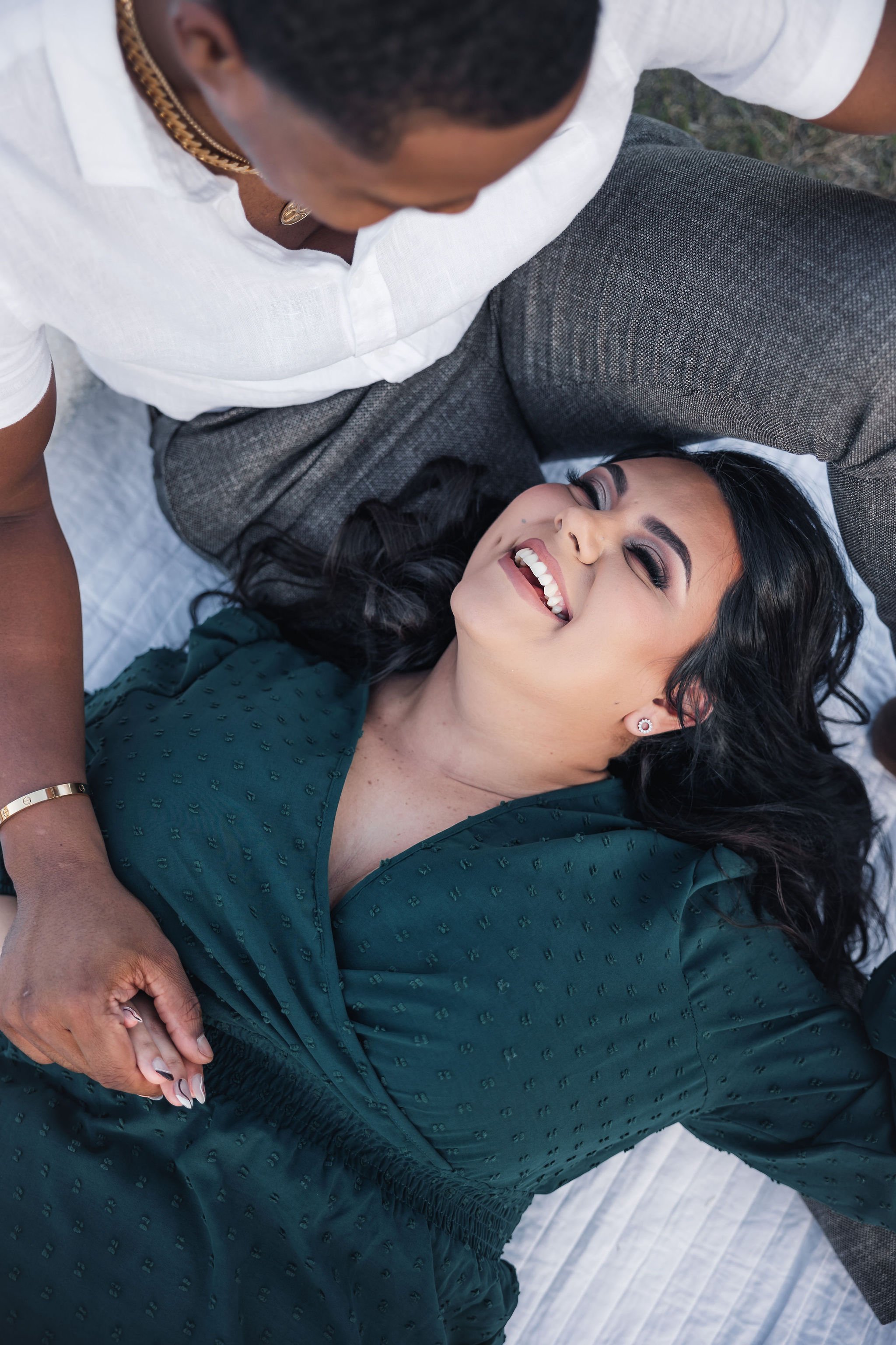A woman lying on a blanket in a green dress, smiling and looking happy, while a man beside her, with his hand holding hers.