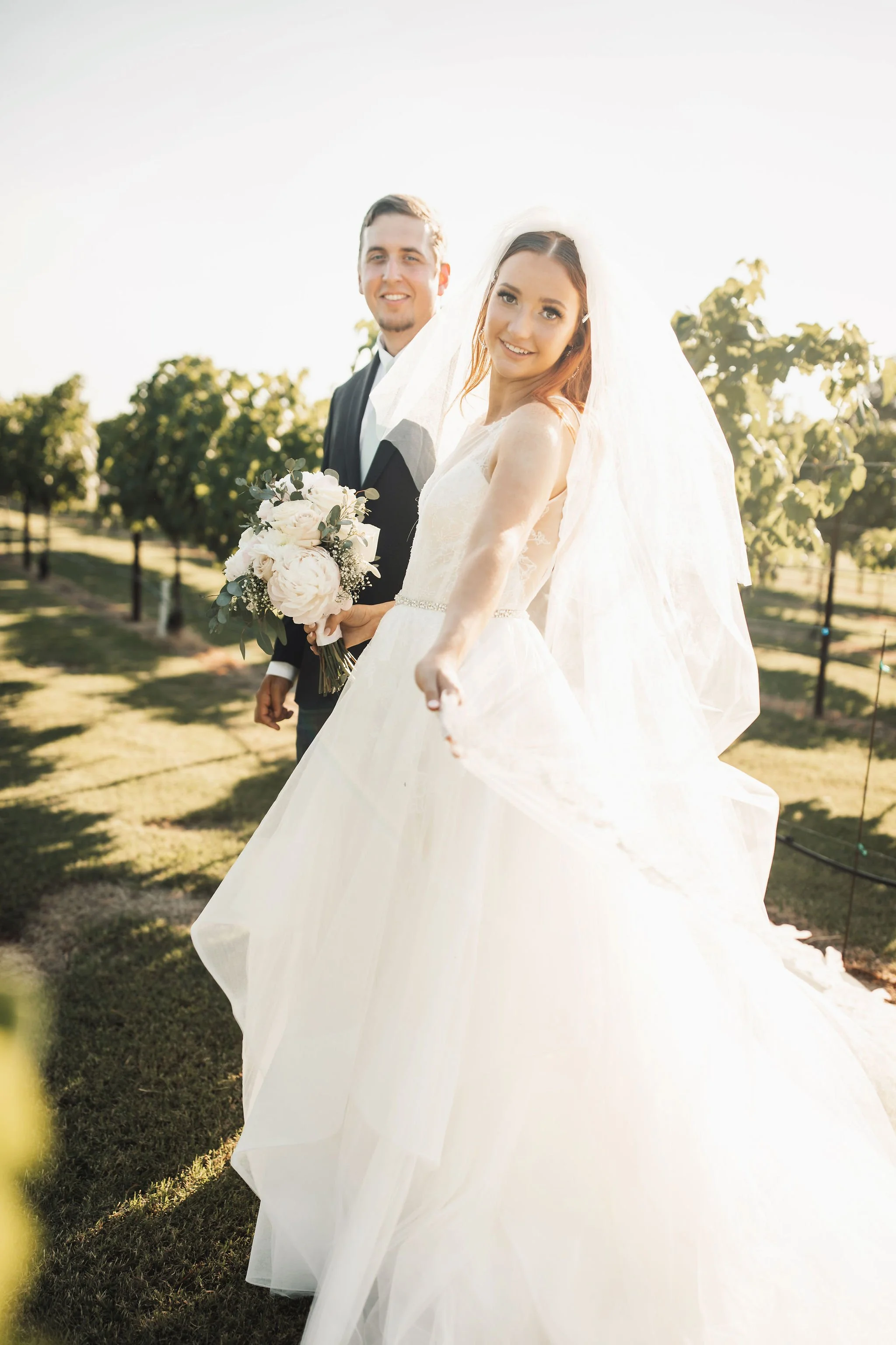 A bride and groom standing outdoors in a vineyard, with the bride holding out her wedding dress and smiling at the camera, while the groom stands behind her holding a bouquet of flowers, both dressed in wedding attire.