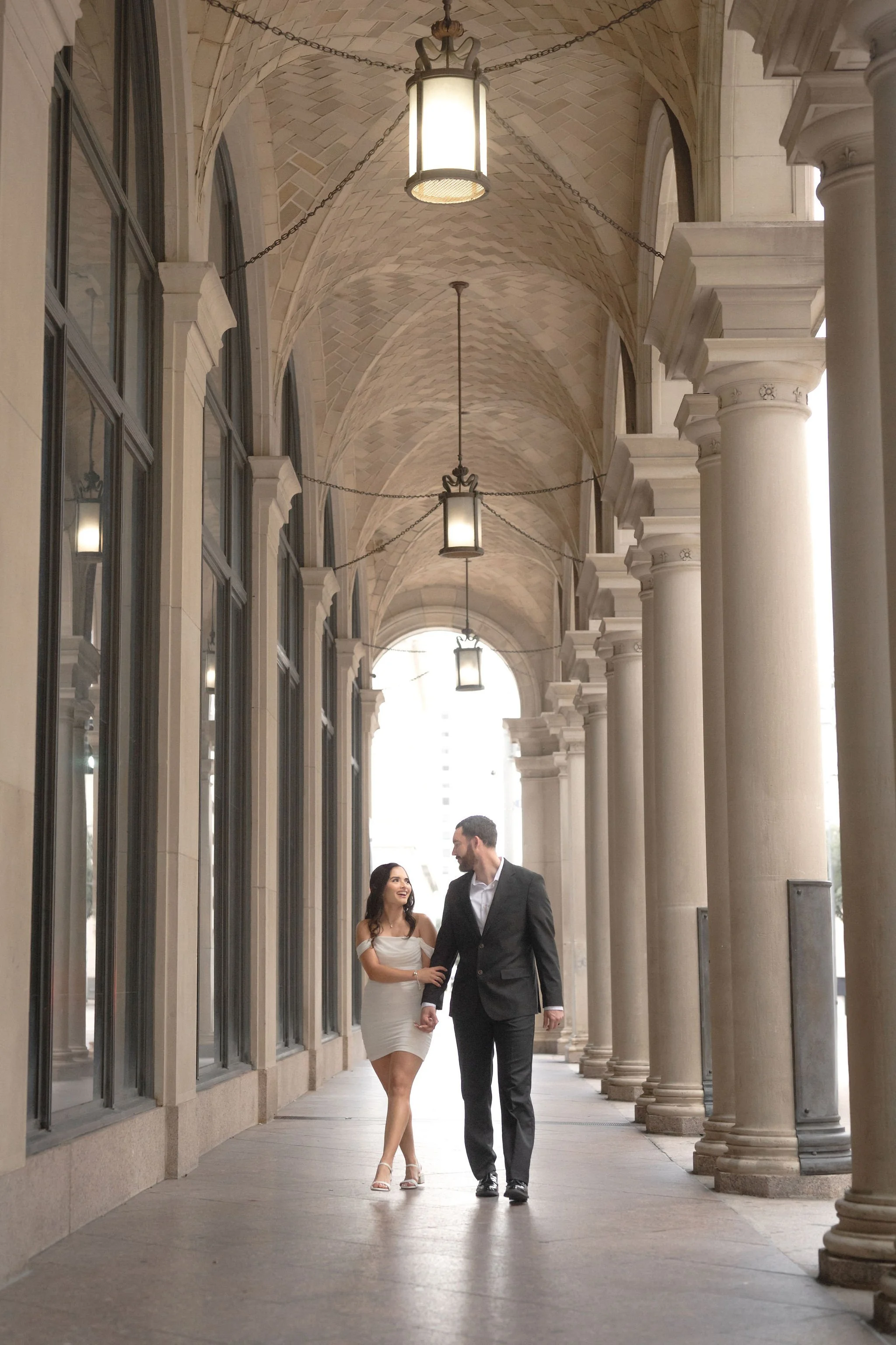 A couple walking hand in hand under an arched walkway with large columns and hanging lanterns, city buildings in the background.