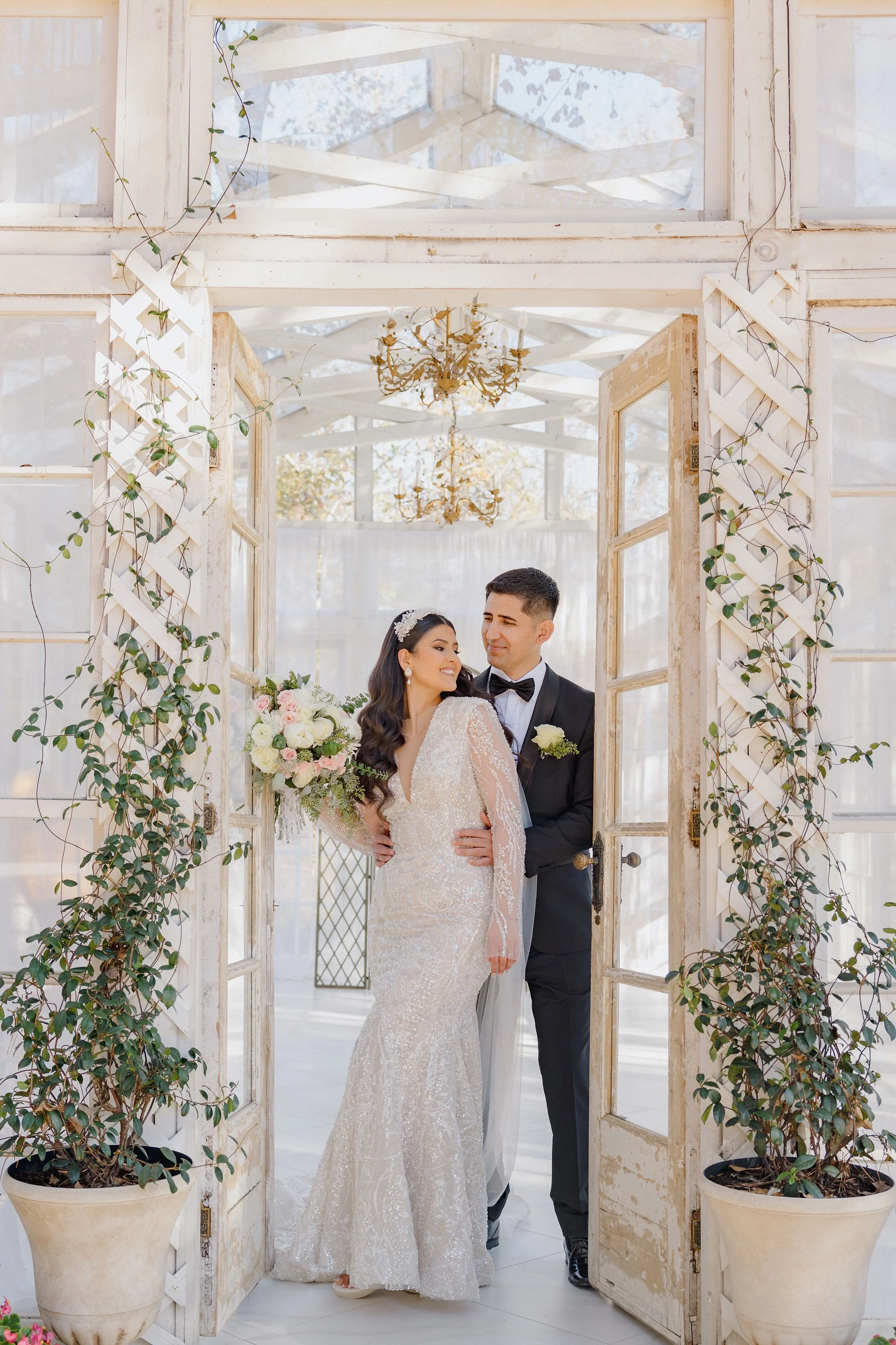 A bride and groom standing in a rustic, vintage-style doorway with open wooden doors and greenery. The bride is holding a bouquet of white and pink flowers and is wearing a long, elegant lace wedding gown. The groom is dressed in a black tuxedo with 