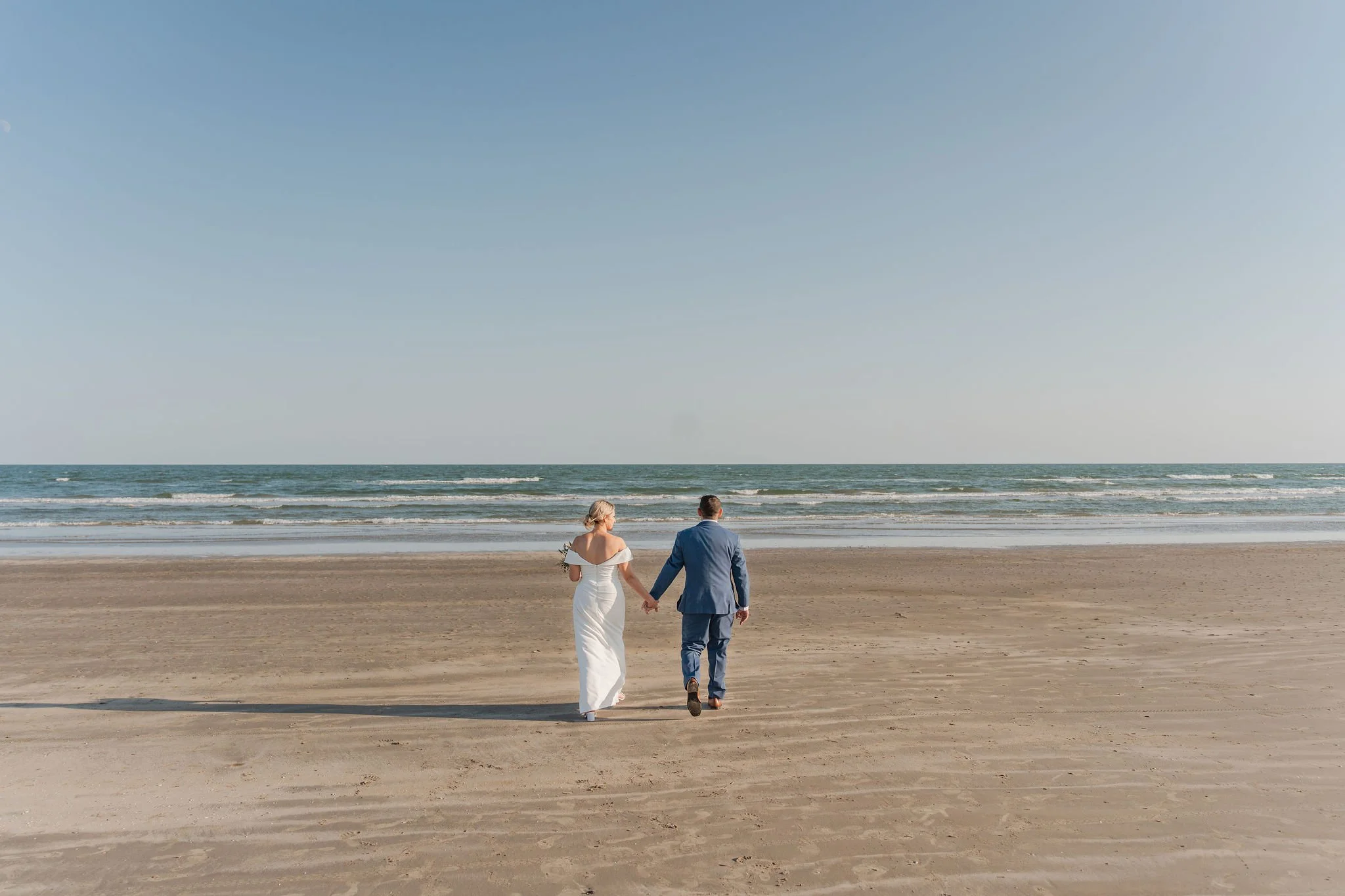 A couple dressed in wedding attire walking hand in hand on a sandy beach towards the ocean.
